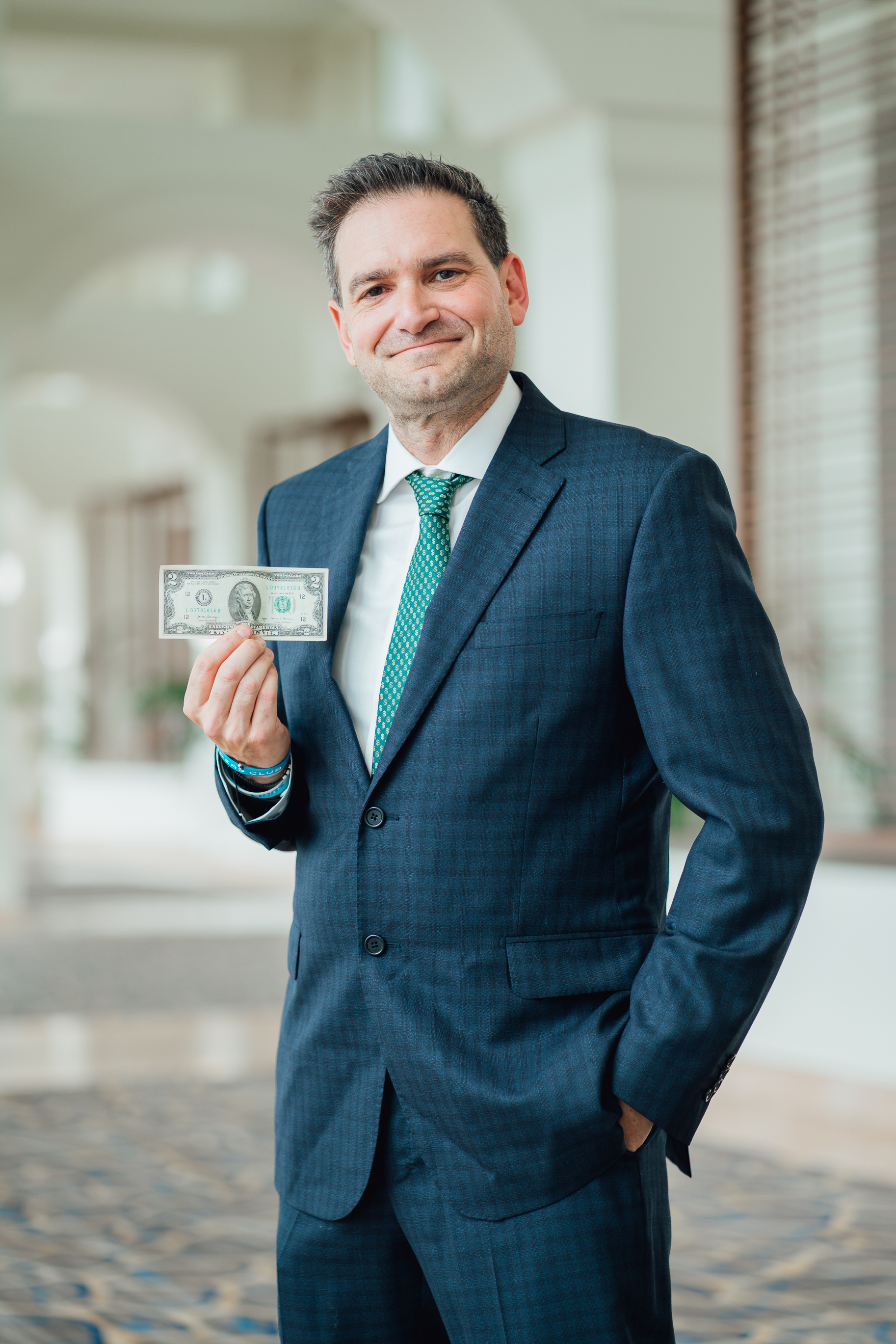 Brad Holcman in a navy suit and green tie holding up a real two dollar bill