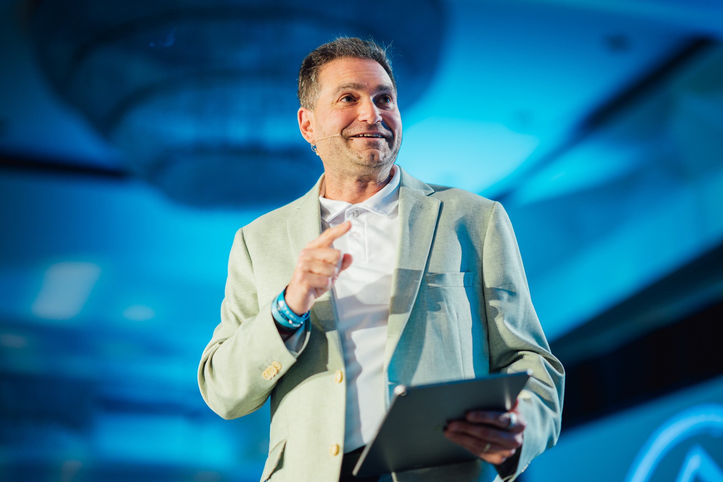 Brad Holcman speaking on stage in a sage green blazer, pointing toward the audience under blue stage lighting